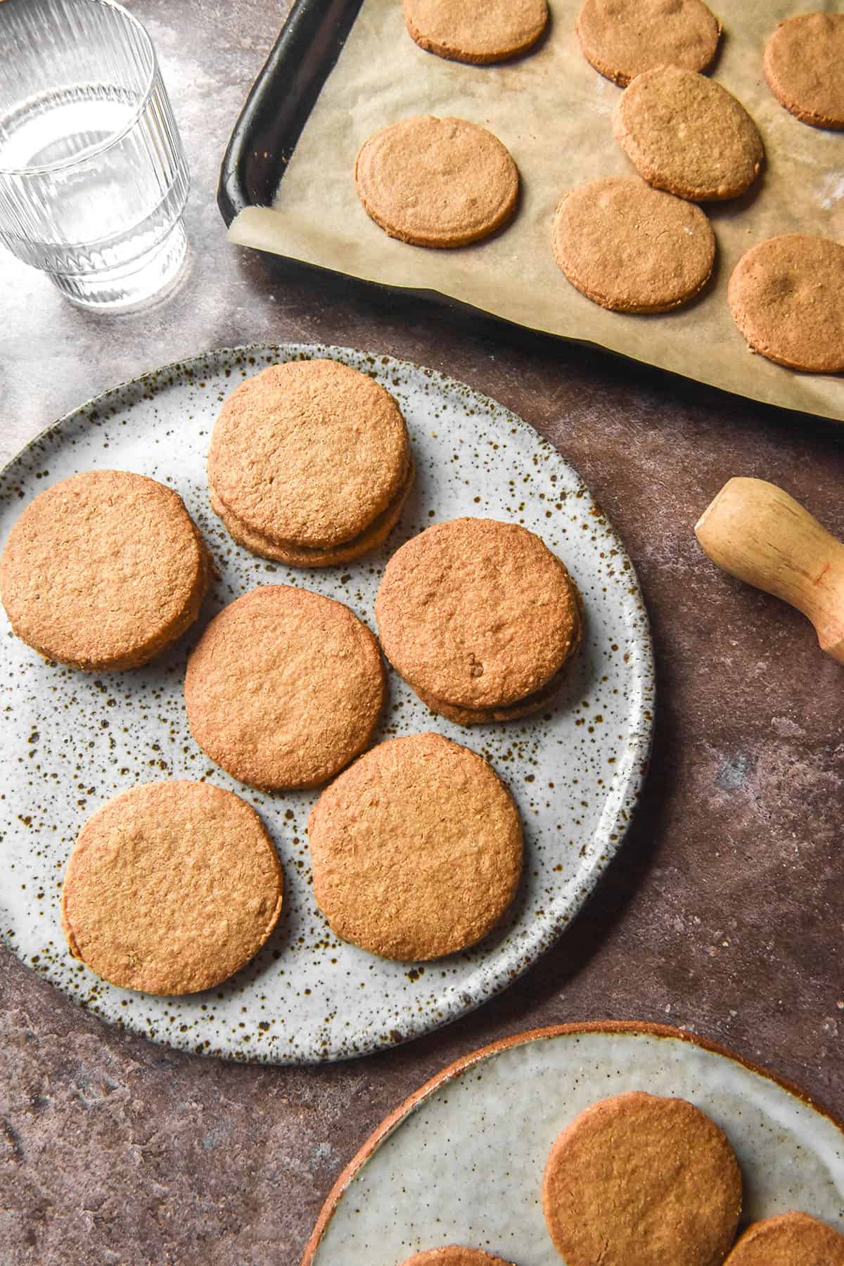 An aerial image of gluten free digestive biscuits on two white ceramic plates and a baking tray atop a medium brown backdrop. A glass of water sits to the left of the biscuits and a rolling pin sits to the bottom right. 