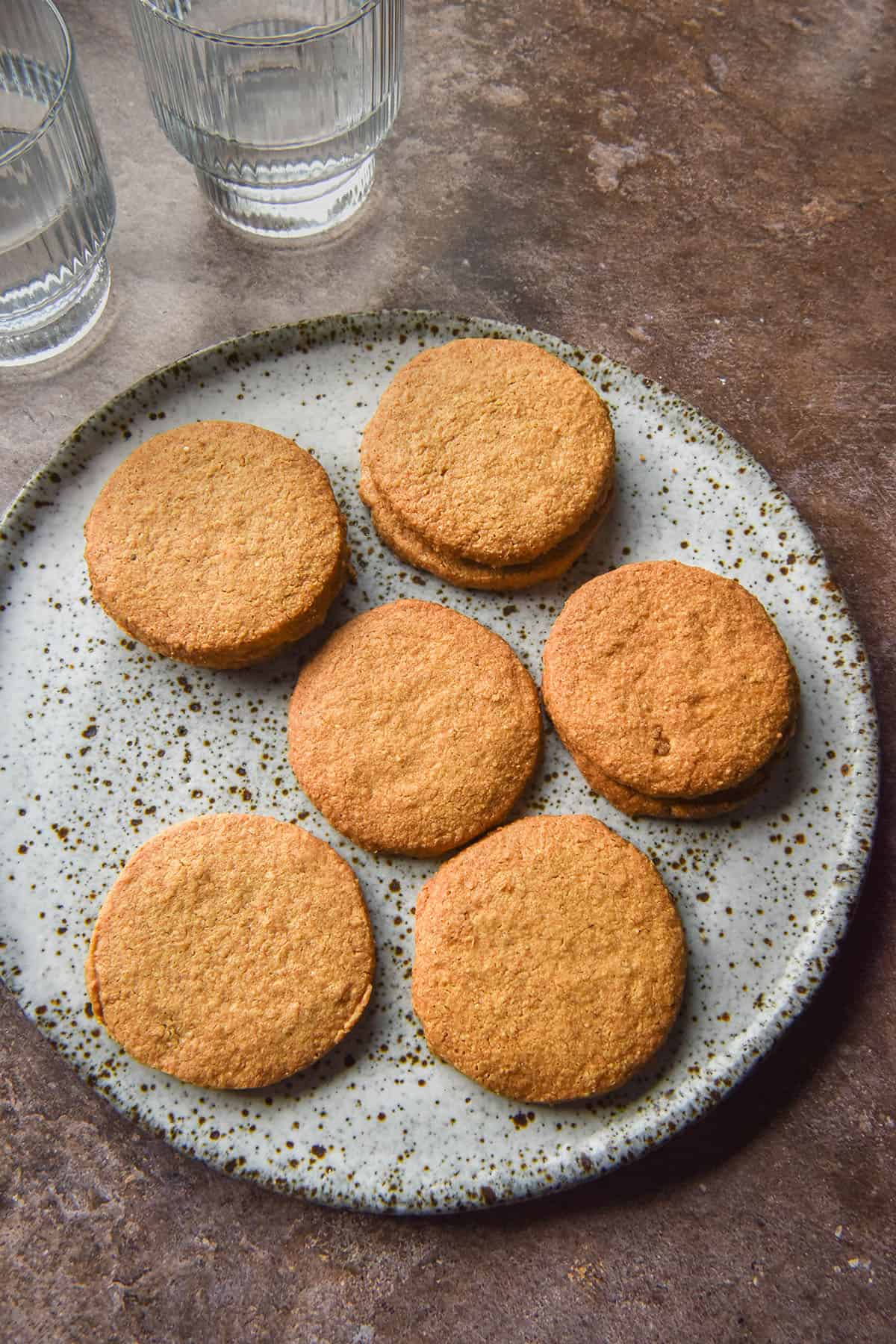 An aerial image of a white speckled ceramic plate topped with gluten free digestive biscuits. The plate sits atop a medium brown backdrop and two glasses of water sit in the top left of the image