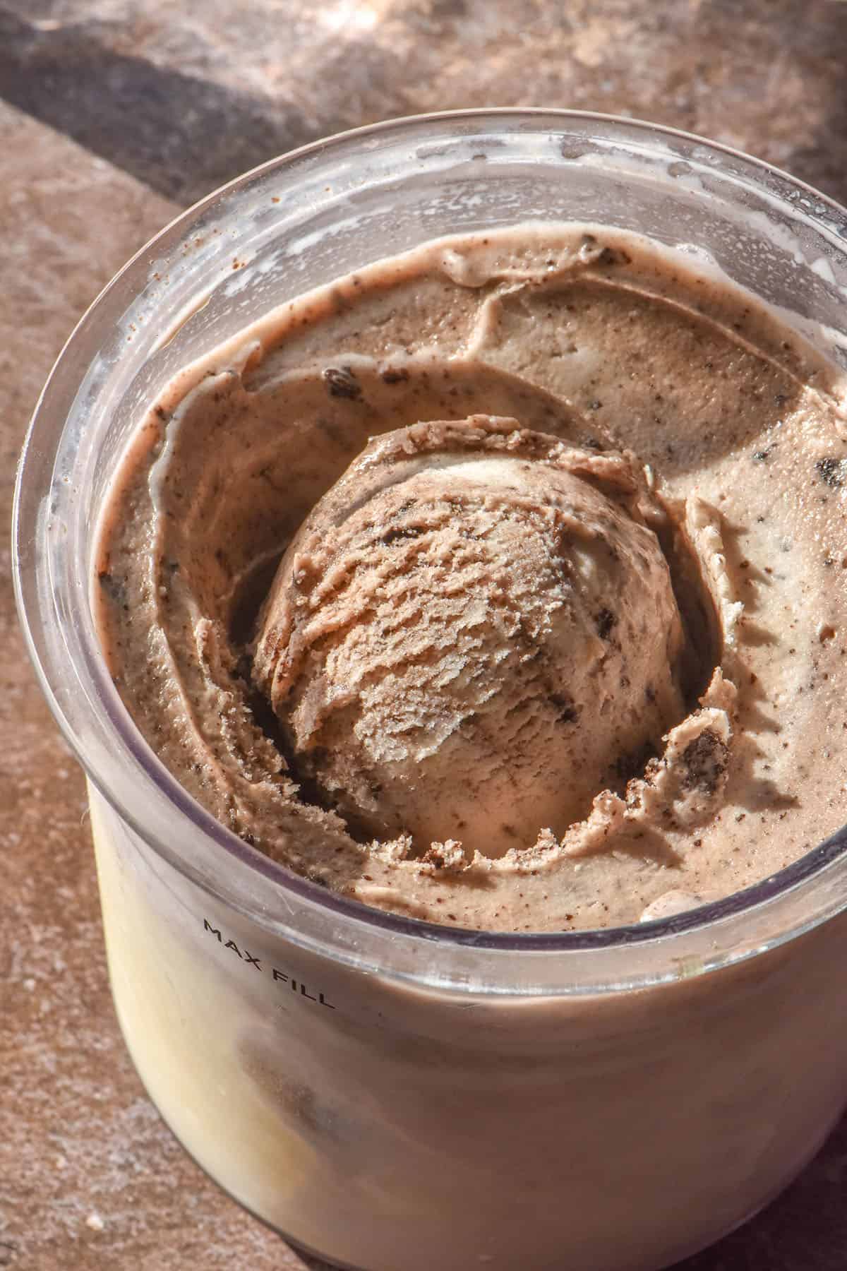 A sunlight close up image of a tub of high protein cookies and cream Ninja Creami ice cream. The tub sits atop a medium brown backdrop with two sunlit glasses of water in the background