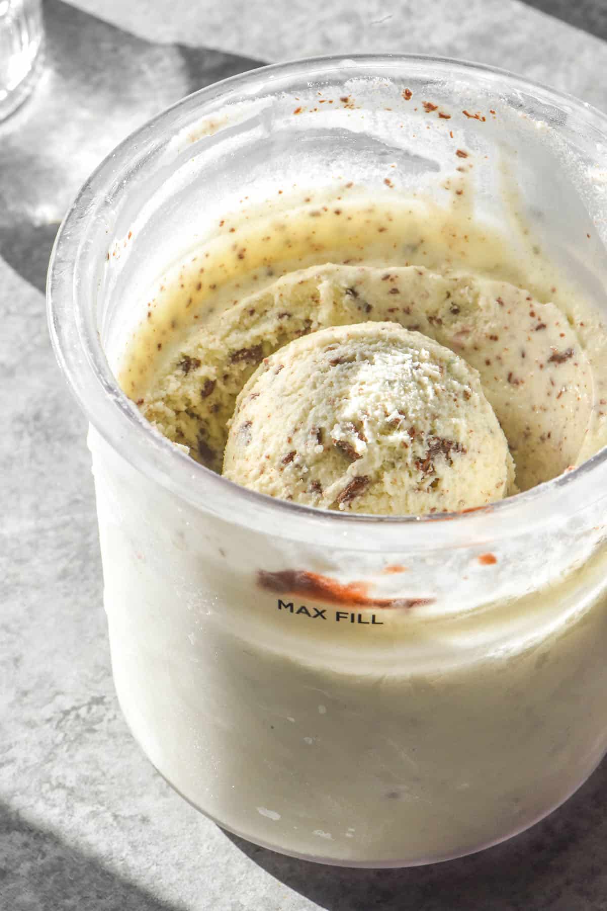 A sunlit close up image of a tub of high protein Ninja Creami mint chocolate chip ice cream. The ice cream sits on a light grey backdrop with sunlit glasses of water in the background