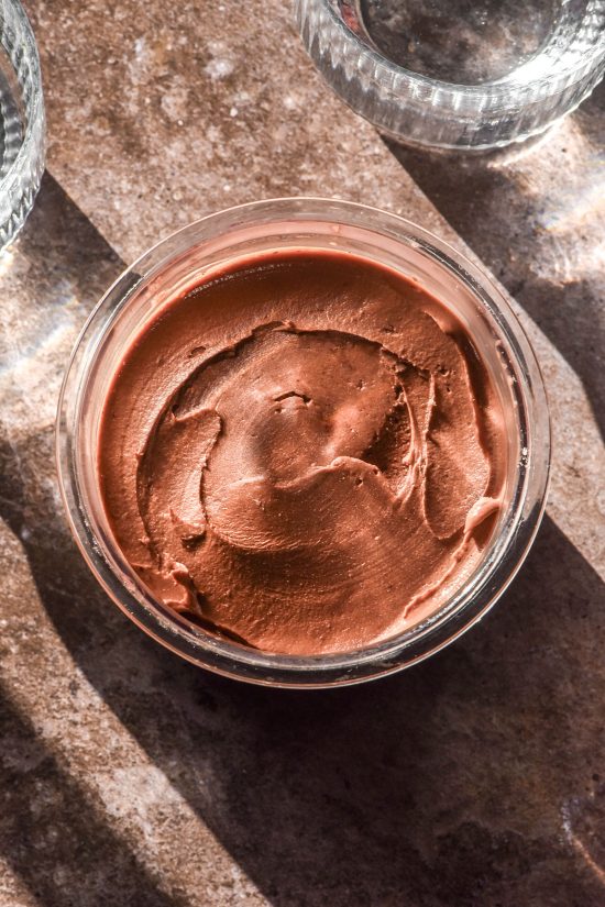 An aerial image of a tub of high protein Ninja creami mocha ice cream on a light brown backdrop. Two sunlit glasses of water sit either side of the ice cream tub