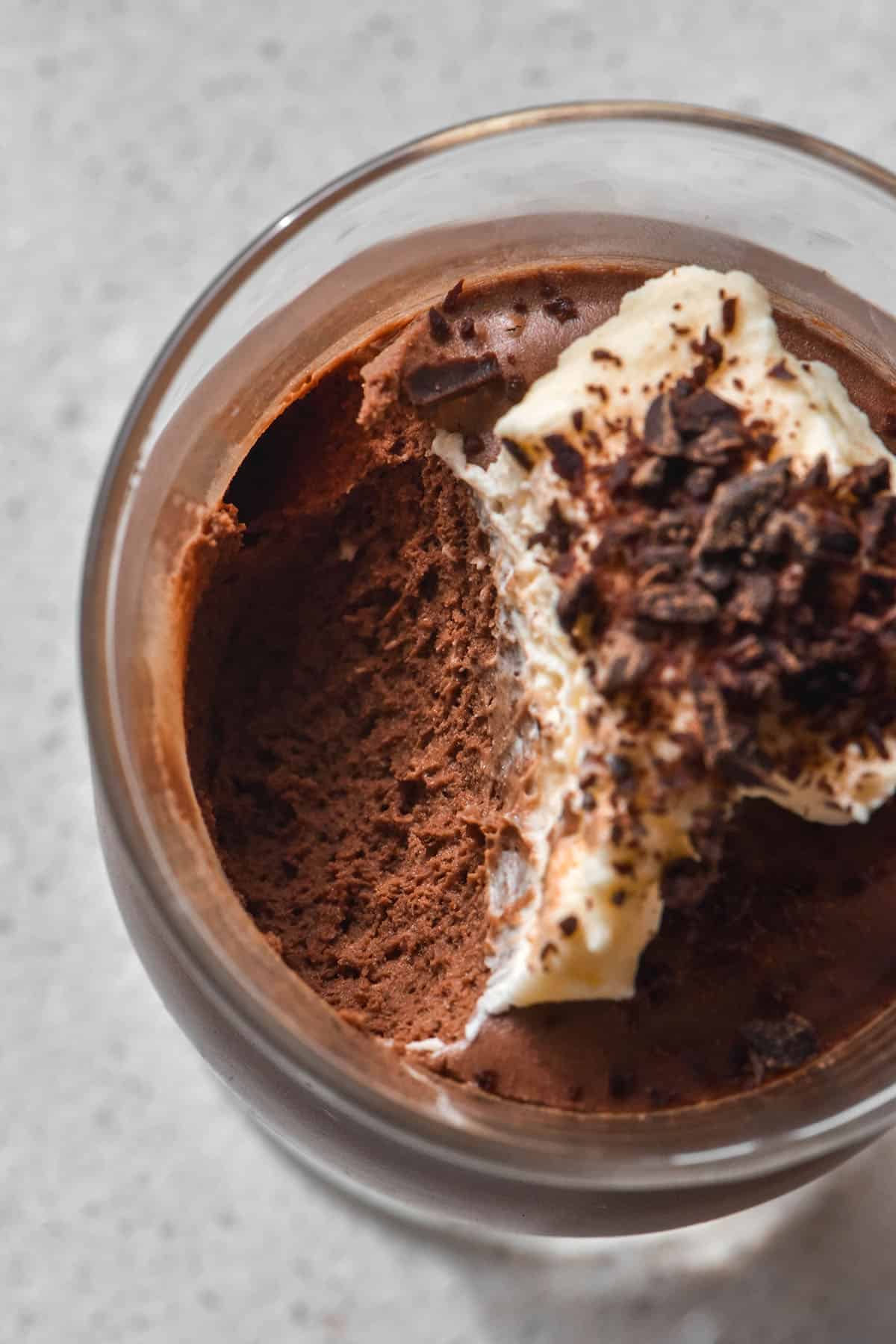 An aerial image of a glass of lactose free chocolate mousse topped with whipped cream and chocolate shavings. A spoonful has been taken, revealing the mousse texture.