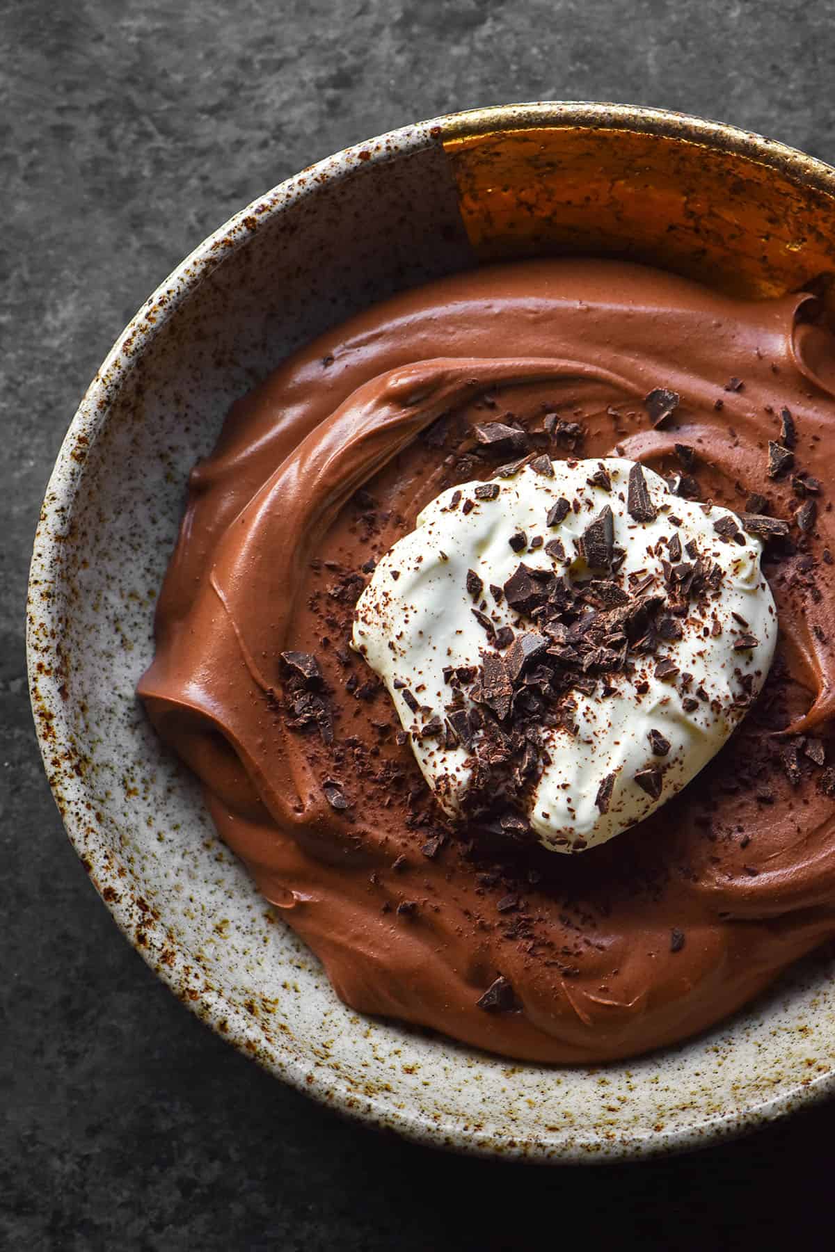 An aerial image of a beige speckled ceramic bowl on a dark grey backdrop filled with lactose free chocolate mousse. The mousse is topped with whipped cream and chocolate shavings.