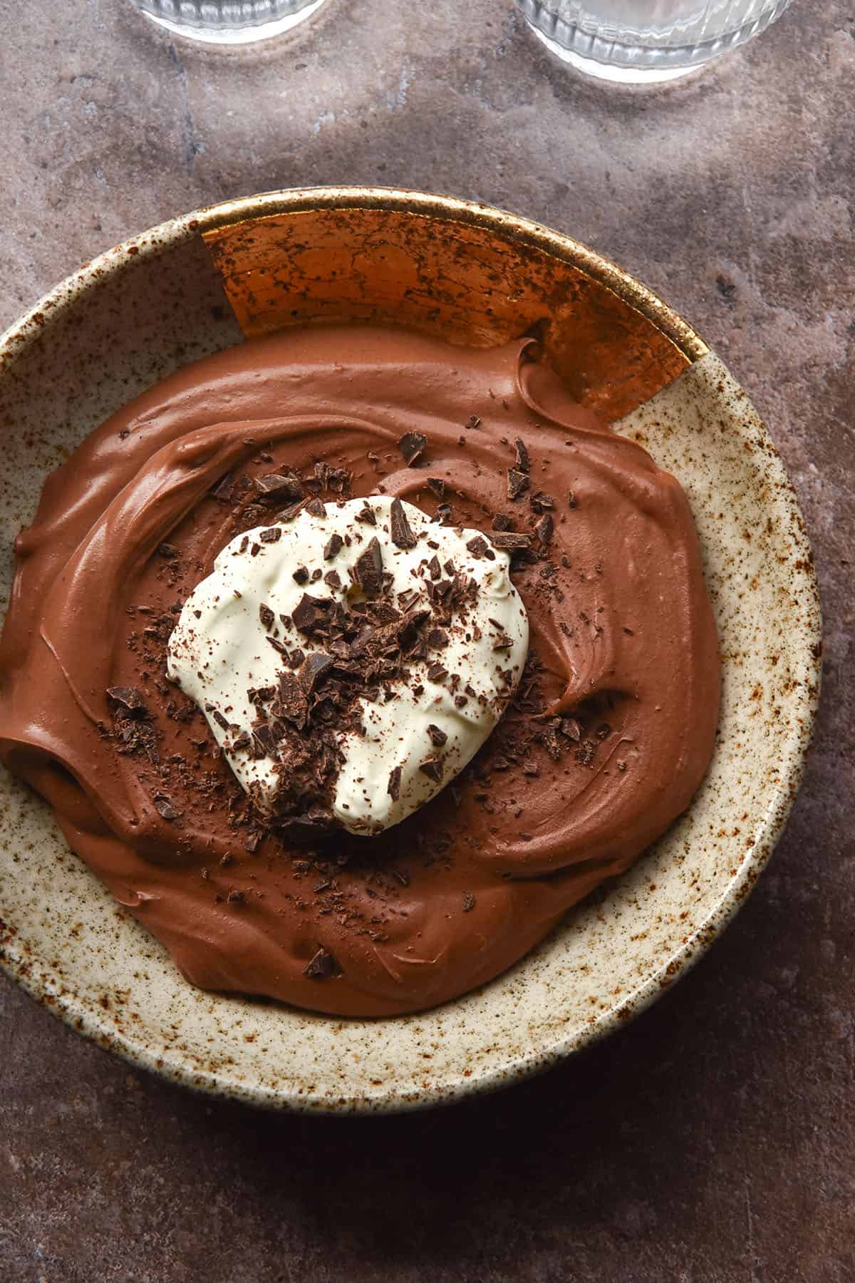 An aerial image of a bowl filled with chocolate mousse that is topped with whipping cream and chocolate shavings. The bowl sits on a dark grey backdrop and two glasses of water sit in the top of the image