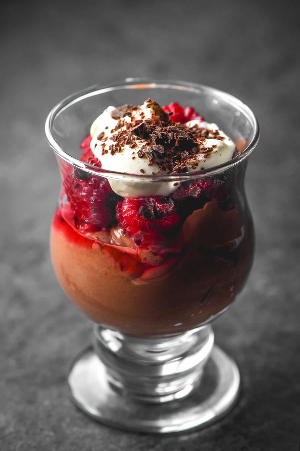 A side on image of a serving glass filled with chocolate mousse, raspberries, whipped cream and chocolate shavings