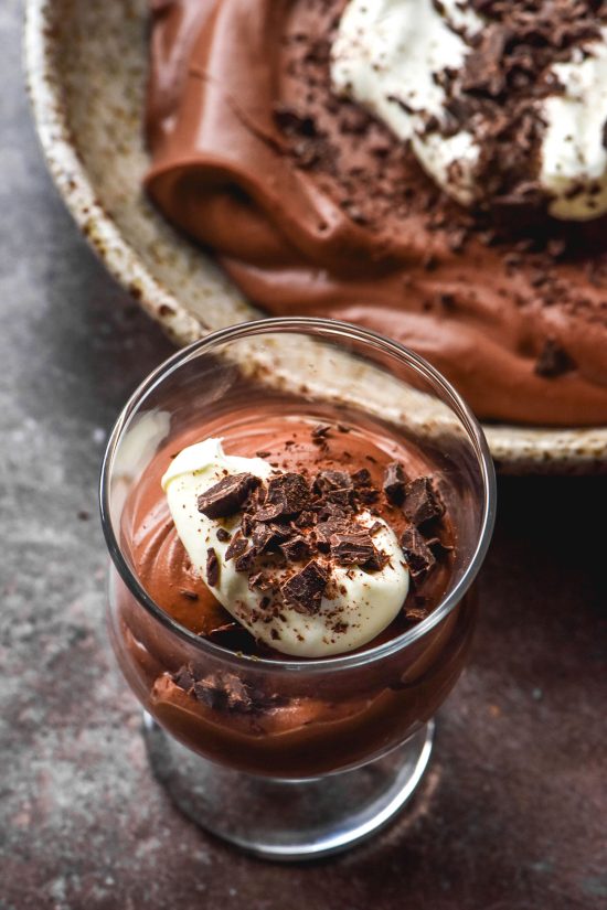 An aerial image of a glass and a bowl filled with lactose free chocolate mousse atop a dark steel backdrop. The mousse is topped with whipped cream and chocolate shavings