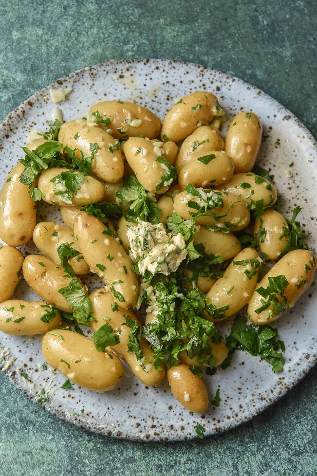 An aerial image of kipfler potatoes covered in low FODMAP garlic butter and parsley. The potatoes sit on a white speckled ceramic serving plate atop a dark green backdrop