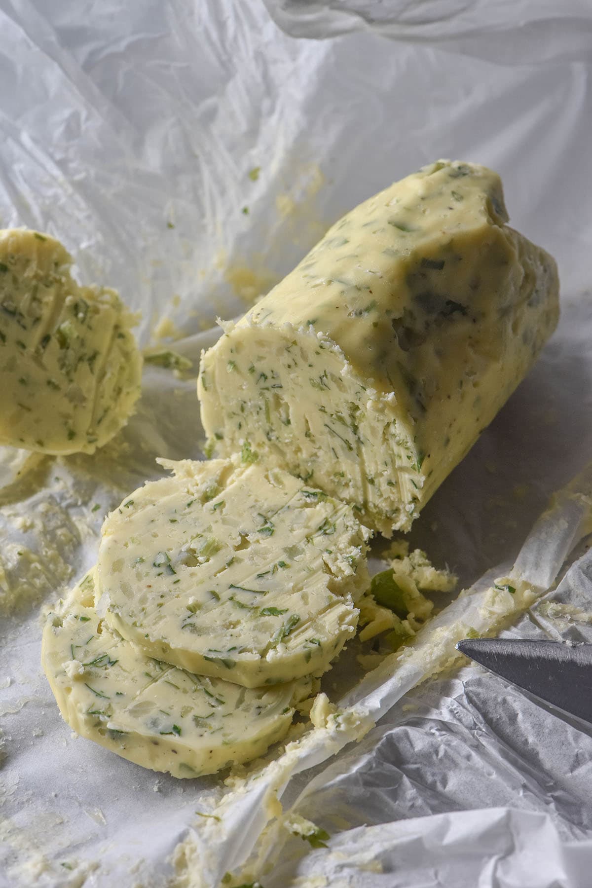 A side on image of a log of low FODMAP garlic butter that has been unwrapped and sits atop the baking paper. The log has been sliced, revealing the garlic and parsley pieces inside