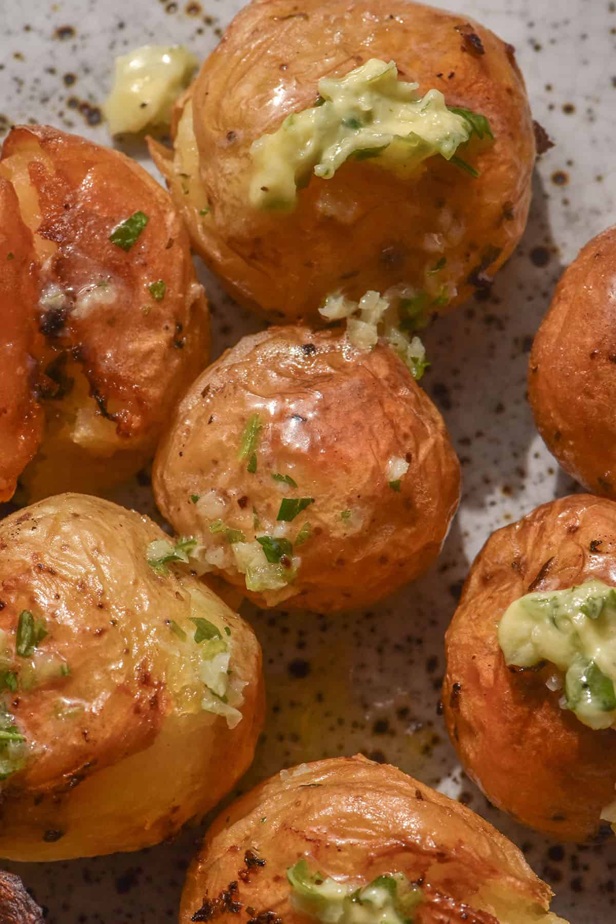 An aerial image of roasted baby potatoes smothered in low FODMAP garlic butter. The potatoes sit atop a white speckled ceramic plate in bright light.