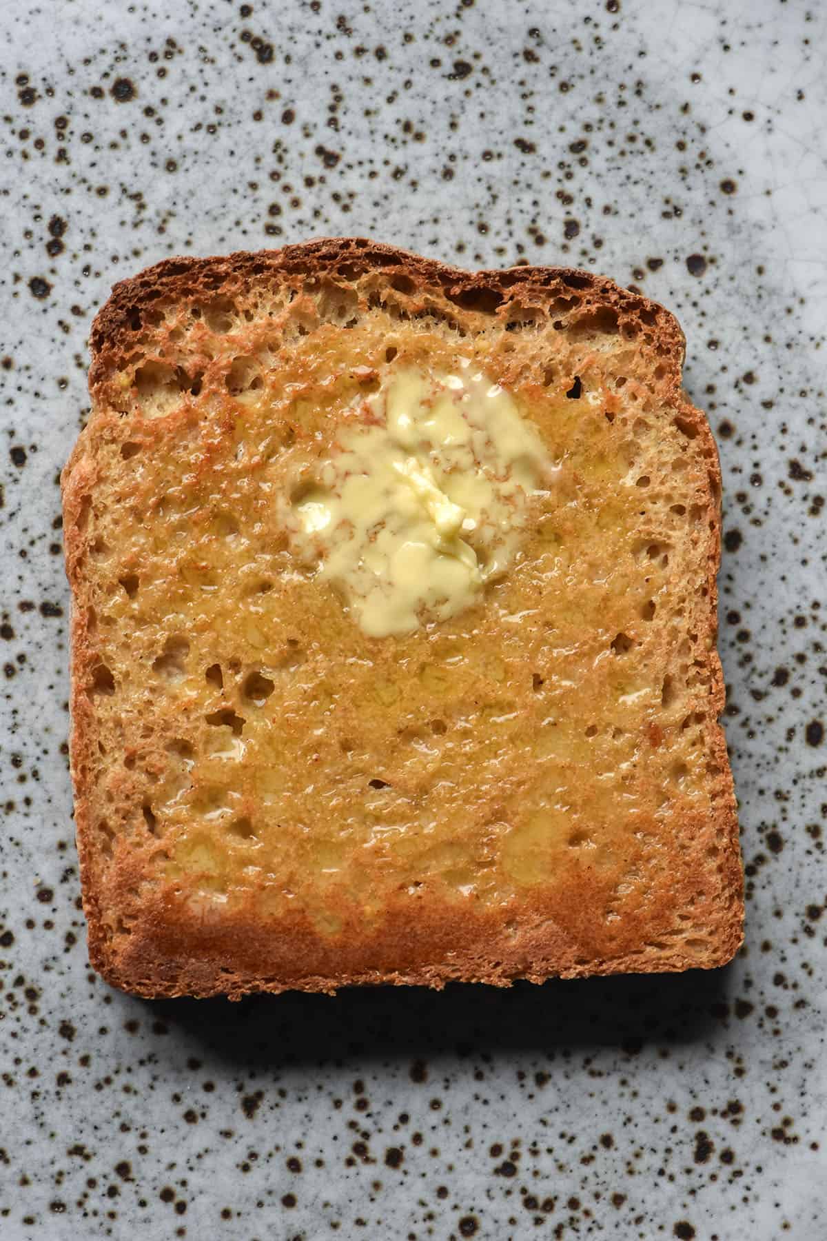 An aerial image of a slice of millet bread toast on a white speckled ceramic plate. The toast has been slathered with butter which has melted into the crumb. 