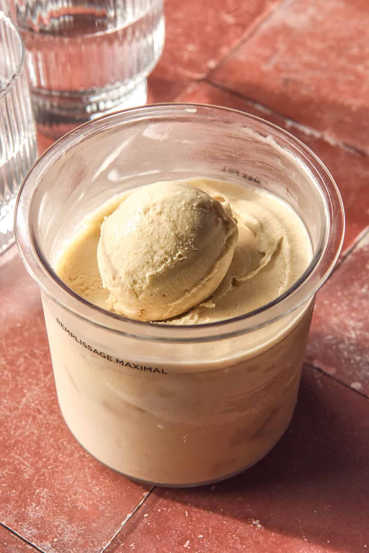 An image of a tub of Ninja Creami banana ice cream on a terracotta tile backdrop. Two sunlit glasses of water sit in the top left of the image.