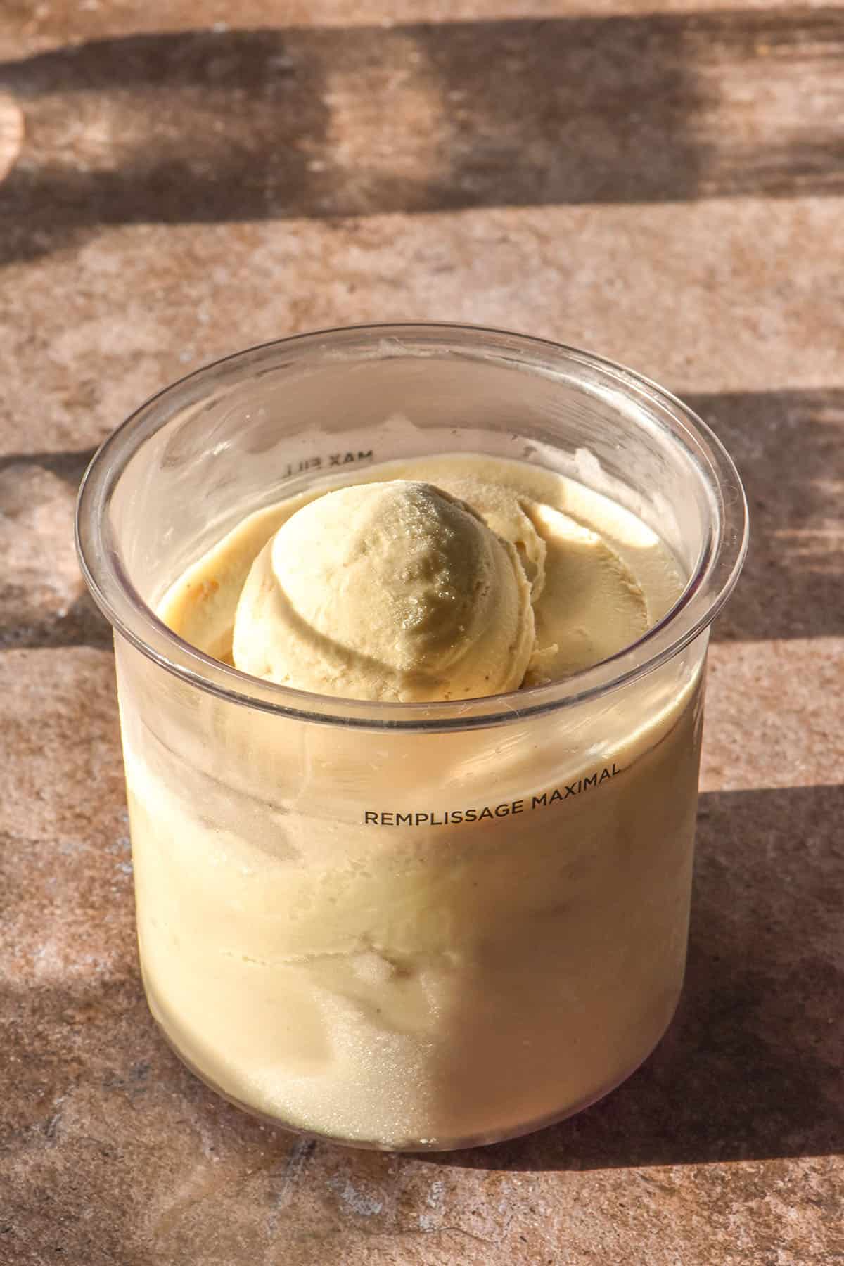 An aerial sunlit image of a tub of Ninja Creami banana ice cream on a light brown backdrop. Two sunlit glasses of water sit in the top left of the image