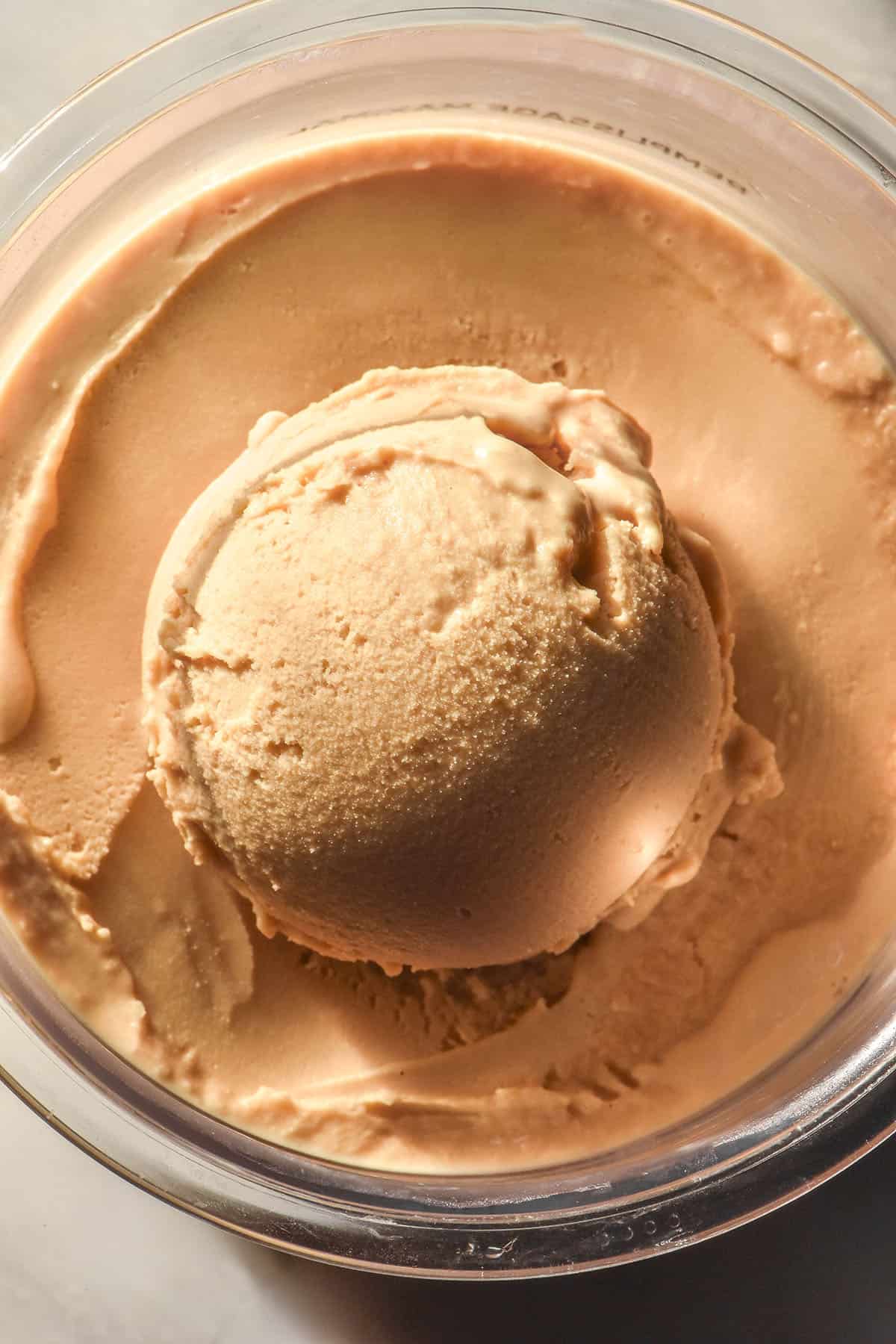 A close up aerial image of a tub of Ninja Creami coffee ice cream on a white marble table. A scoop has been taken from the ice cream and sits perched in the middle of the tub.