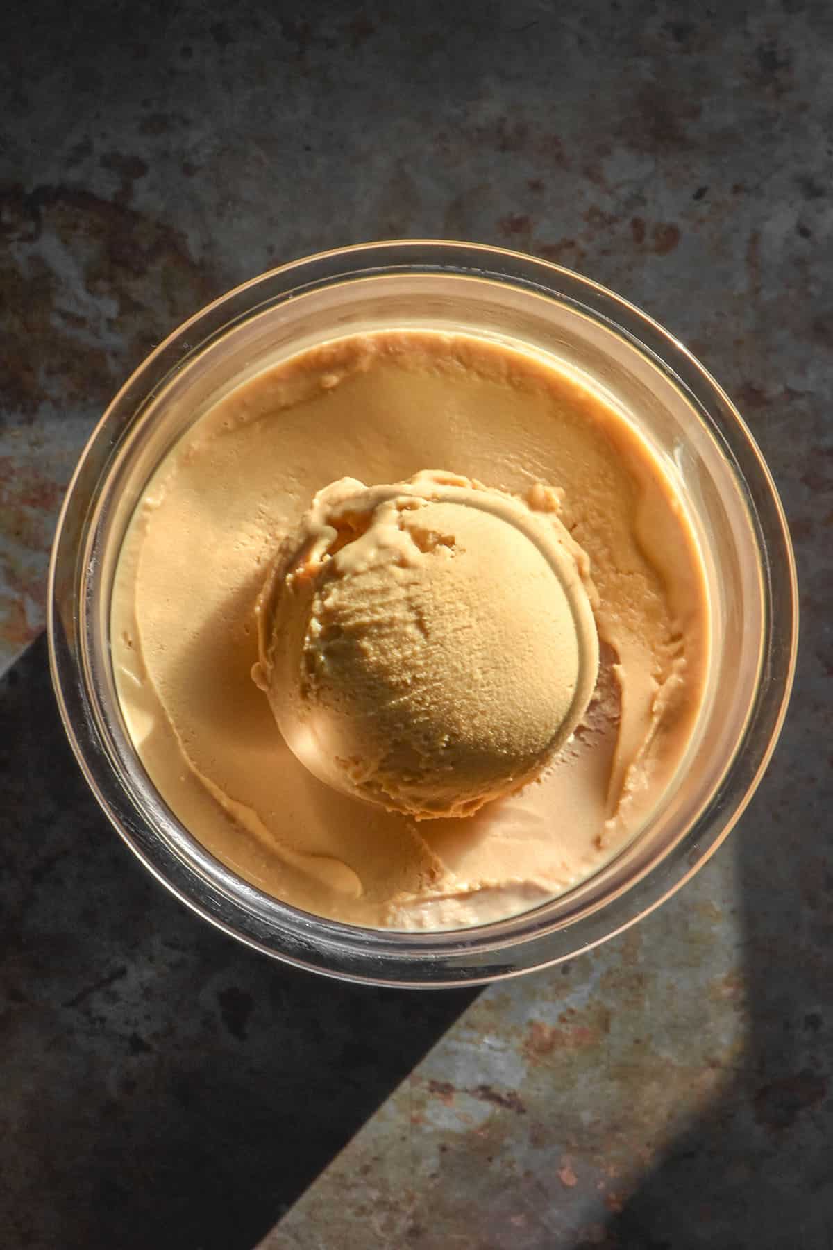 An aerial image of a tub of Ninja Creami coffee ice cream on a dark moody steel backdrop in contrasting sunlight. 