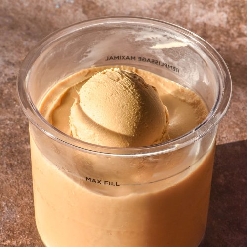 A brightly lit side on image of a tub of Ninja Creami coffee ice cream. The tub sits atop a medium brown backdrop and a scoop of the ice cream is perched in the centre of the tub