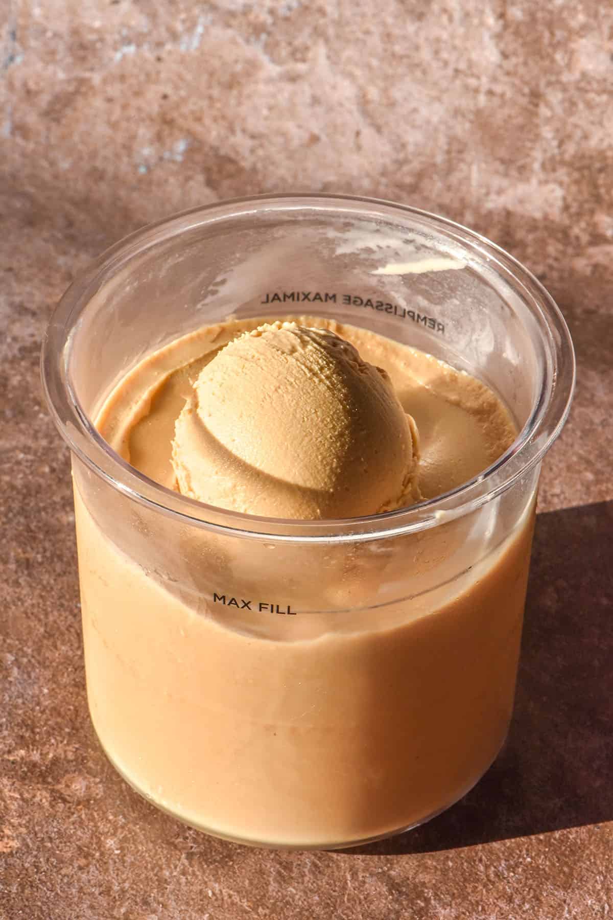 A brightly lit side on image of a tub of Ninja Creami coffee ice cream. The tub sits atop a medium brown backdrop and a scoop of the ice cream is perched in the centre of the tub