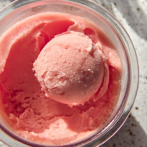 A brightly lit aerial image of a tub of Ninja Creami watermelon sorbet. The tub sits atop a white speckled ceramic plate and sunlit glasses of water sit in the background