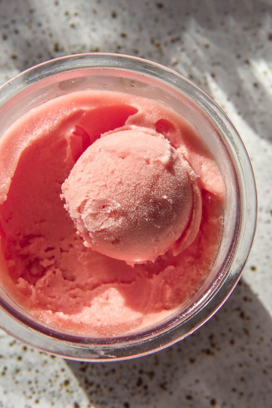 A brightly lit aerial image of a tub of Ninja Creami watermelon sorbet. The tub sits atop a white speckled ceramic plate and sunlit glasses of water sit in the background