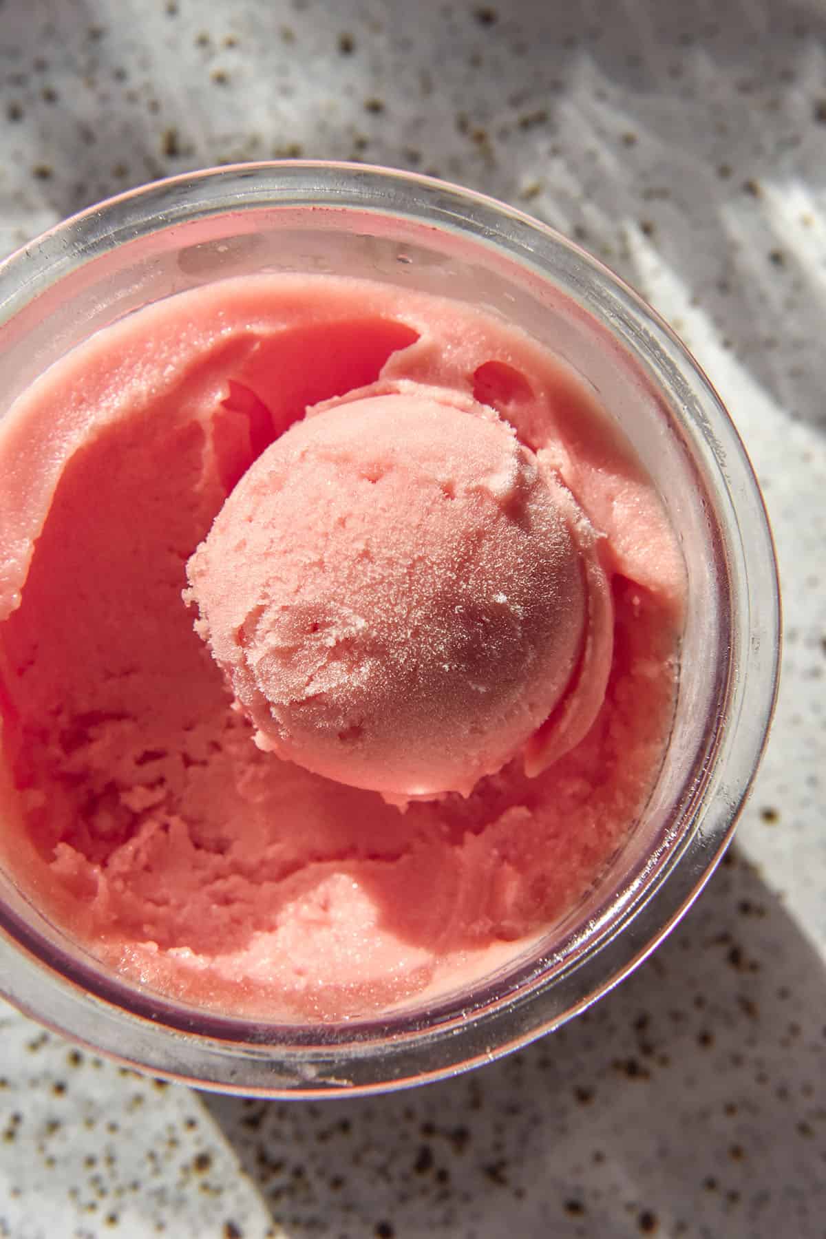 A brightly lit aerial image of a tub of Ninja Creami watermelon sorbet. The tub sits atop a white speckled ceramic plate and sunlit glasses of water sit in the background