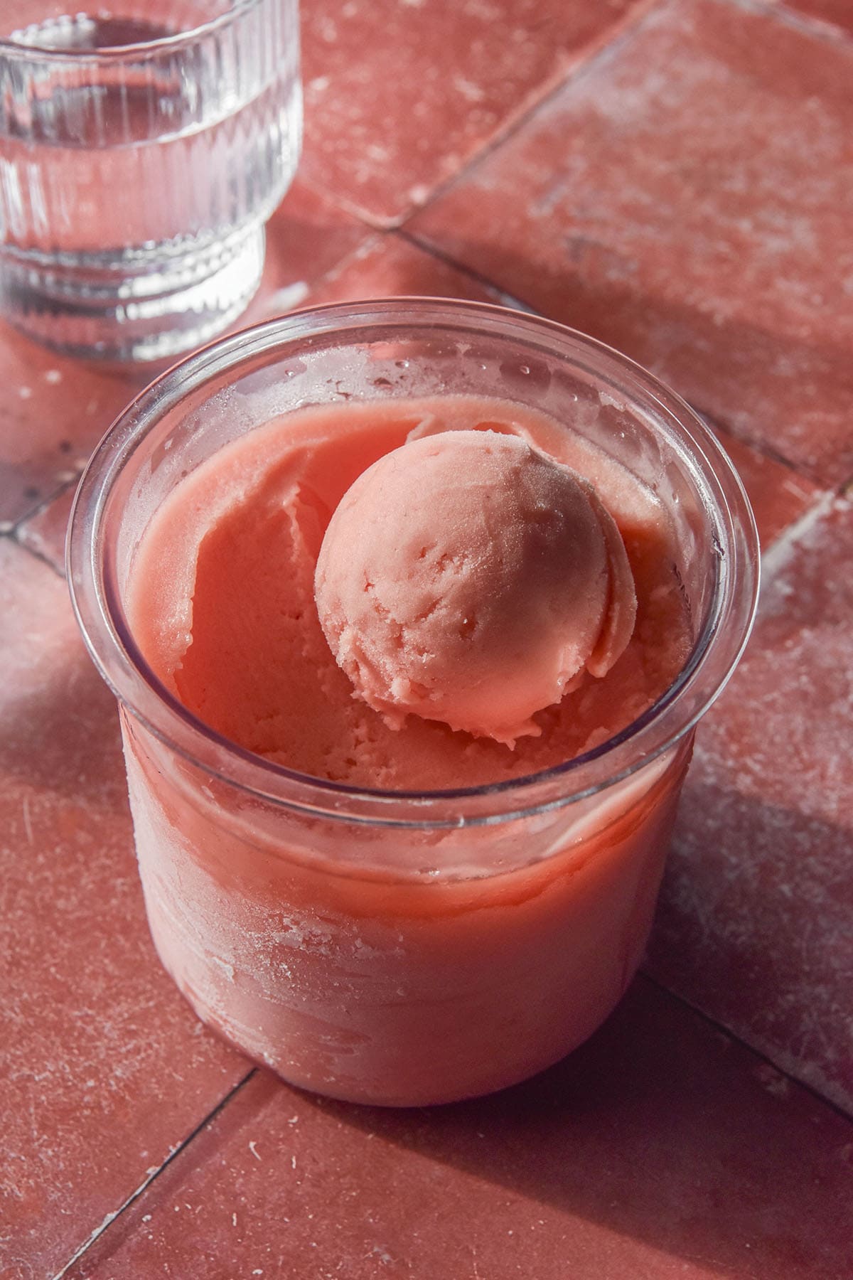 A side on image of a tub of Ninja Creami watermelon sorbet on a terracotta tile backdrop. A sunlit glass of water sits in the top left corner