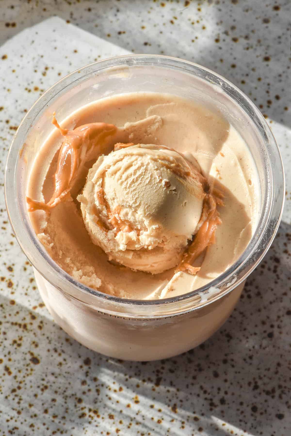 An aerial sunlit image of a tub of high protein peanut butter Ninja Creami on a white speckled ceramic plate