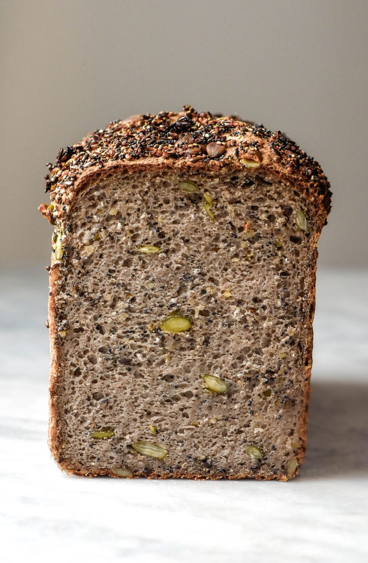 A side on image of a loaf of gluten free seeded buckwheat bread on a white marble table against a white backdrop. the loaf has been sliced to reveal the seeded inner crumb of the bread.