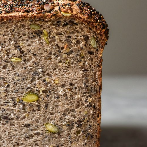 A side on image of a loaf of gluten free seeded buckwheat bread on a white marble table against a white backdrop. the loaf has been sliced to reveal the seeded inner crumb of the bread.