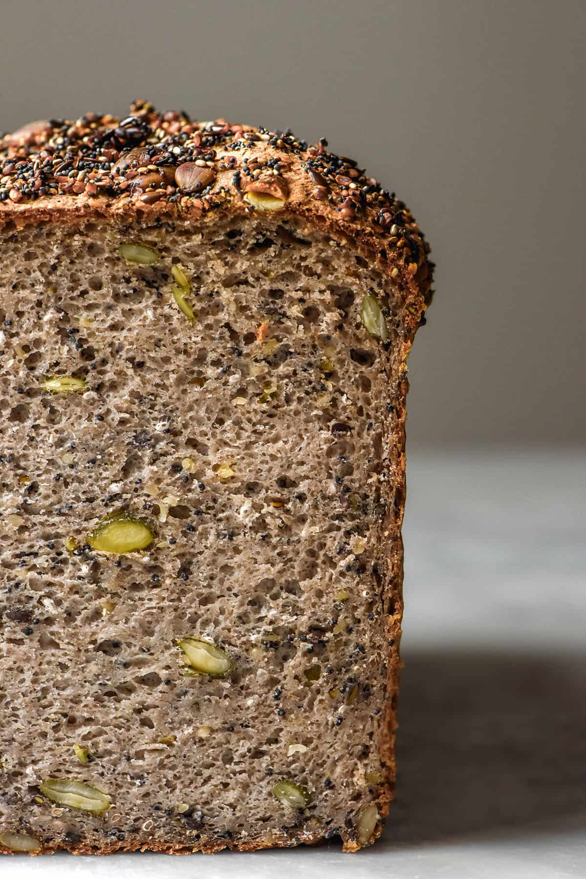 A side on image of a loaf of gluten free seeded buckwheat bread on a white marble table against a white backdrop. the loaf has been sliced to reveal the seeded inner crumb of the bread.