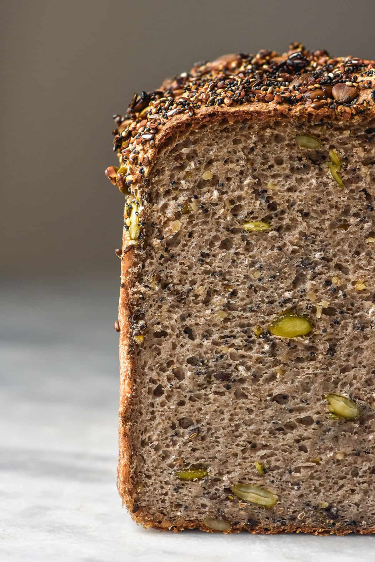 A close up side on image of a loaf of gluten free seeded buckwheat bread on a white marble table against a white backdrop. the loaf has been sliced to reveal the seeded inner crumb of the bread.