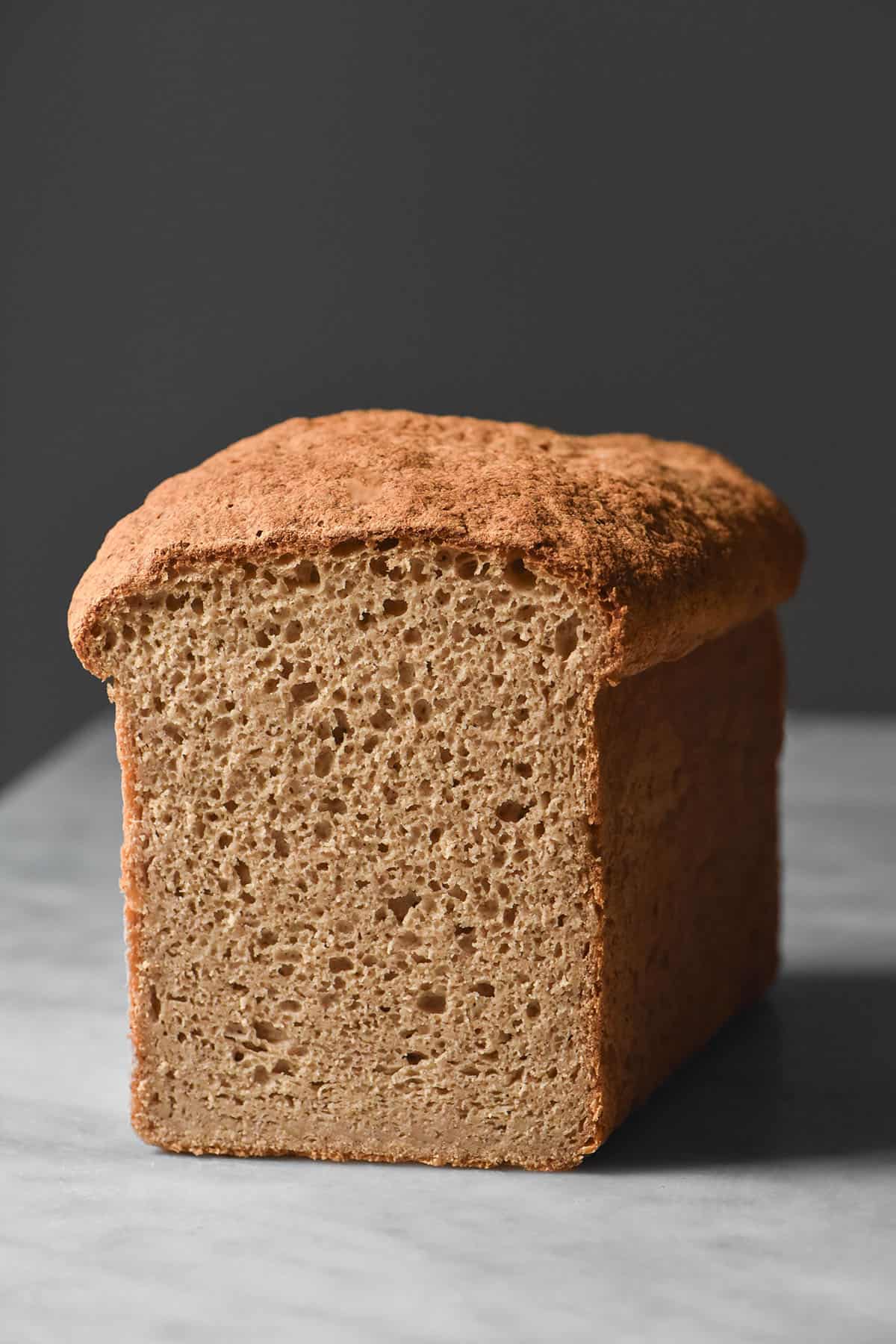 A moody side on image of a loaf of sorghum bread sitting on a marble table against a dark backdrop. The loaf has been sliced to reveal the crumb inside.