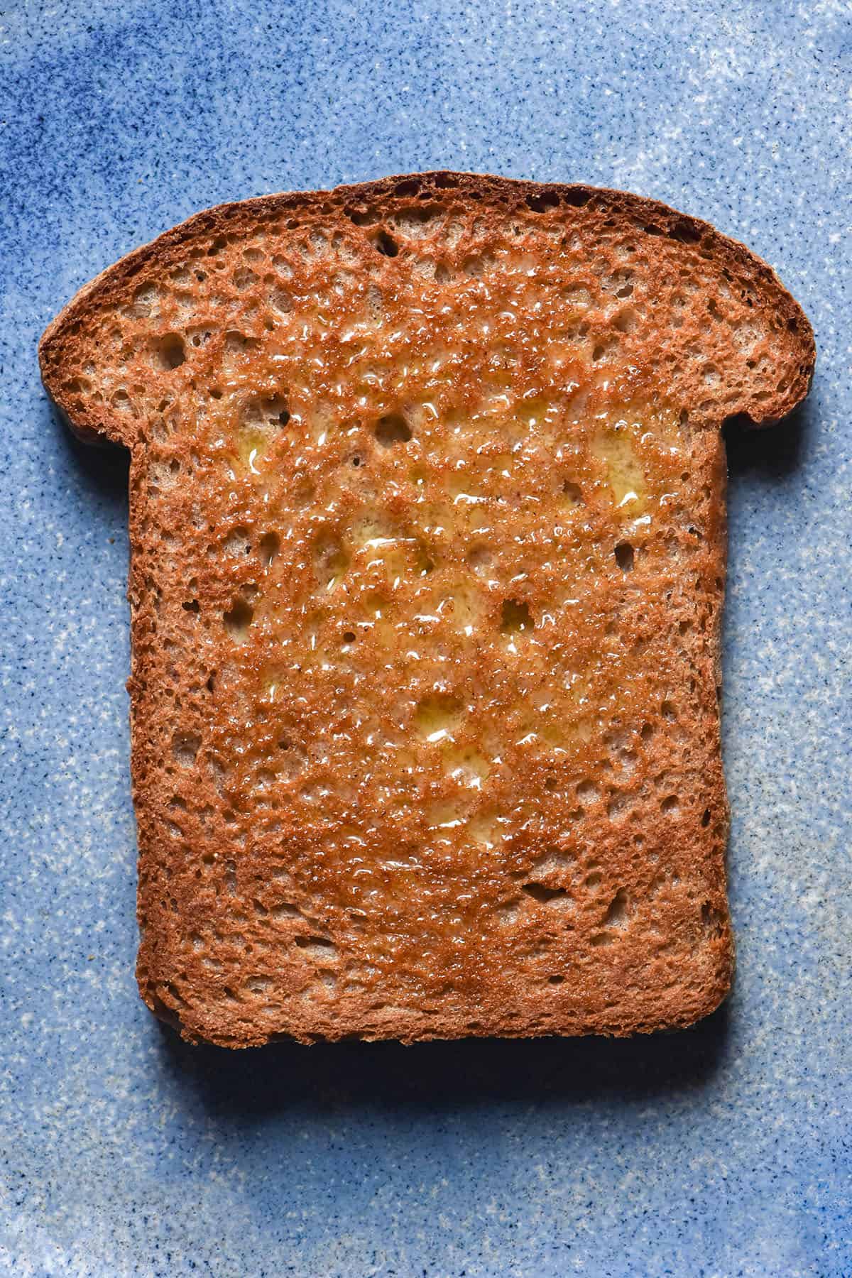 An aerial close up image of a slice of toasted sorghum bread on a light blue ceramic plate. The bread is golden brown and slathered with melted butter