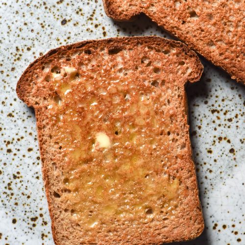 An aerial image of two slices of sorghum bread toast that are slathered in butter and casually arranged on a white speckled ceramic plate