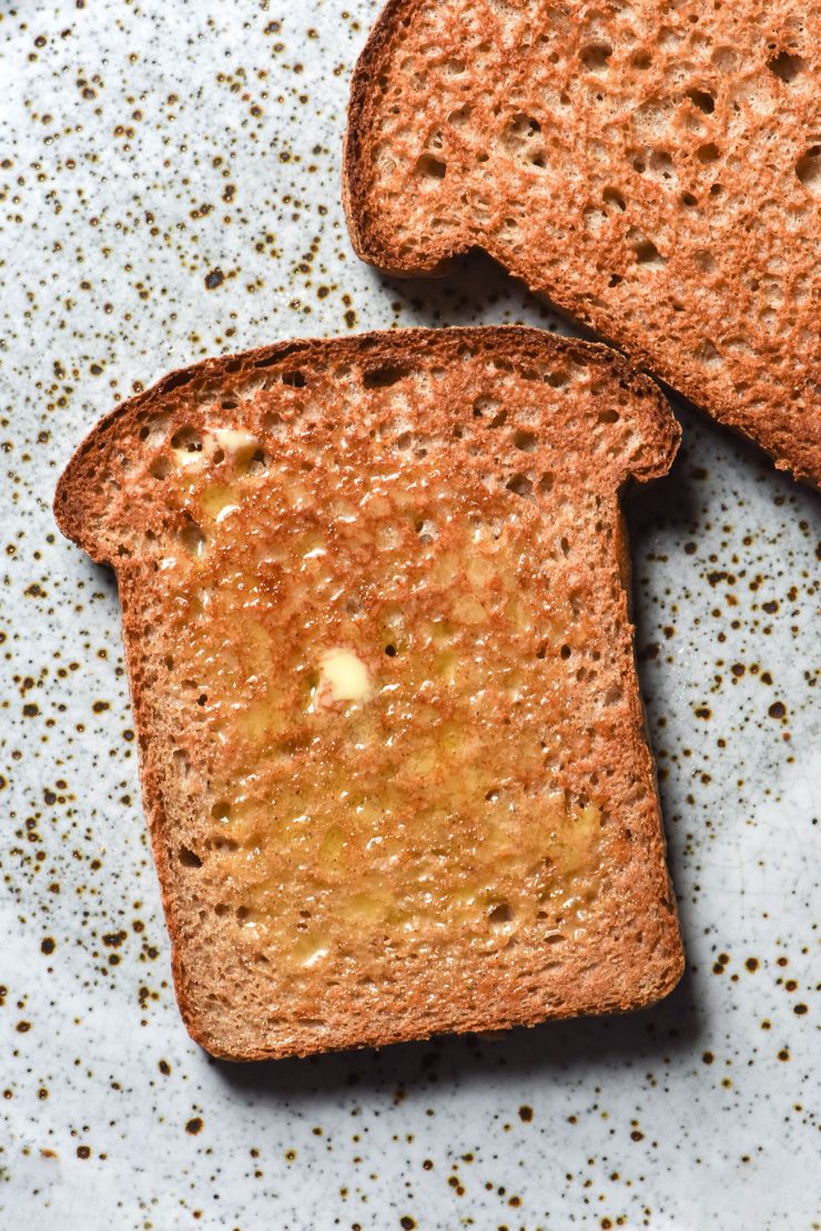 An aerial image of two slices of sorghum bread toast that are slathered in butter and casually arranged on a white speckled ceramic plate