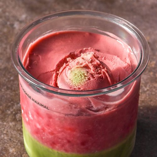 A side on image of a strawberry matcha Ninja Creami on a dark grey backdrop. A scoop of the ice cream sits atop the tub