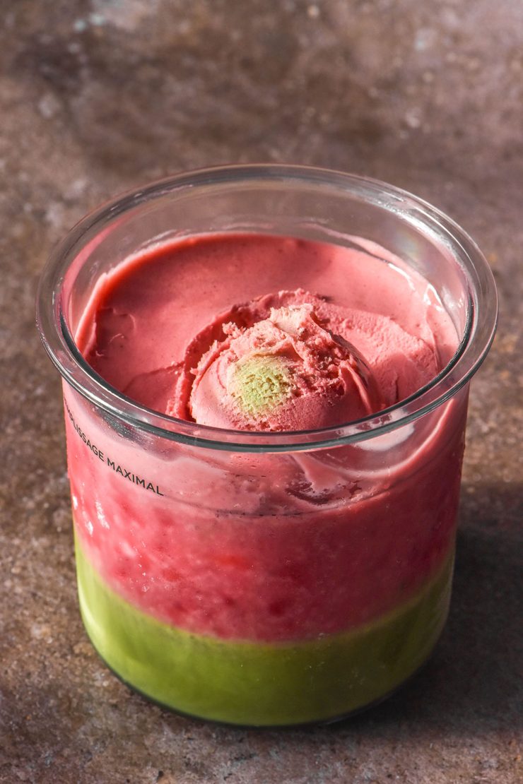 A side on image of a strawberry matcha Ninja Creami on a dark grey backdrop. A scoop of the ice cream sits atop the tub