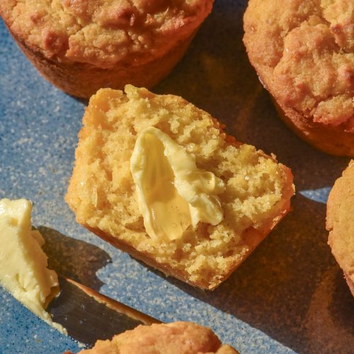 An aerial image of gluten free cornbread muffins on a light blue ceramic plate. The central cornbread muffin has been split and slathered in butter and honey.