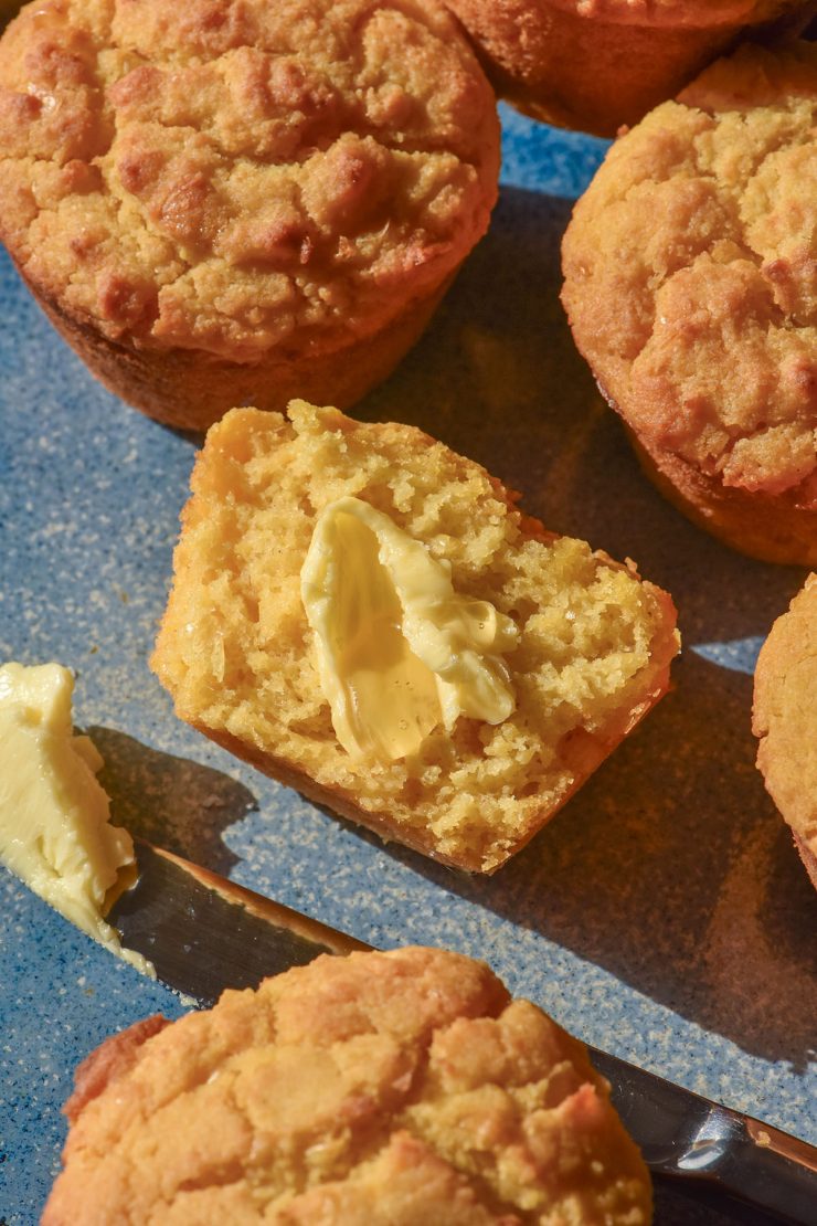 An aerial image of gluten free cornbread muffins on a light blue ceramic plate. The central cornbread muffin has been split and slathered in butter and honey.