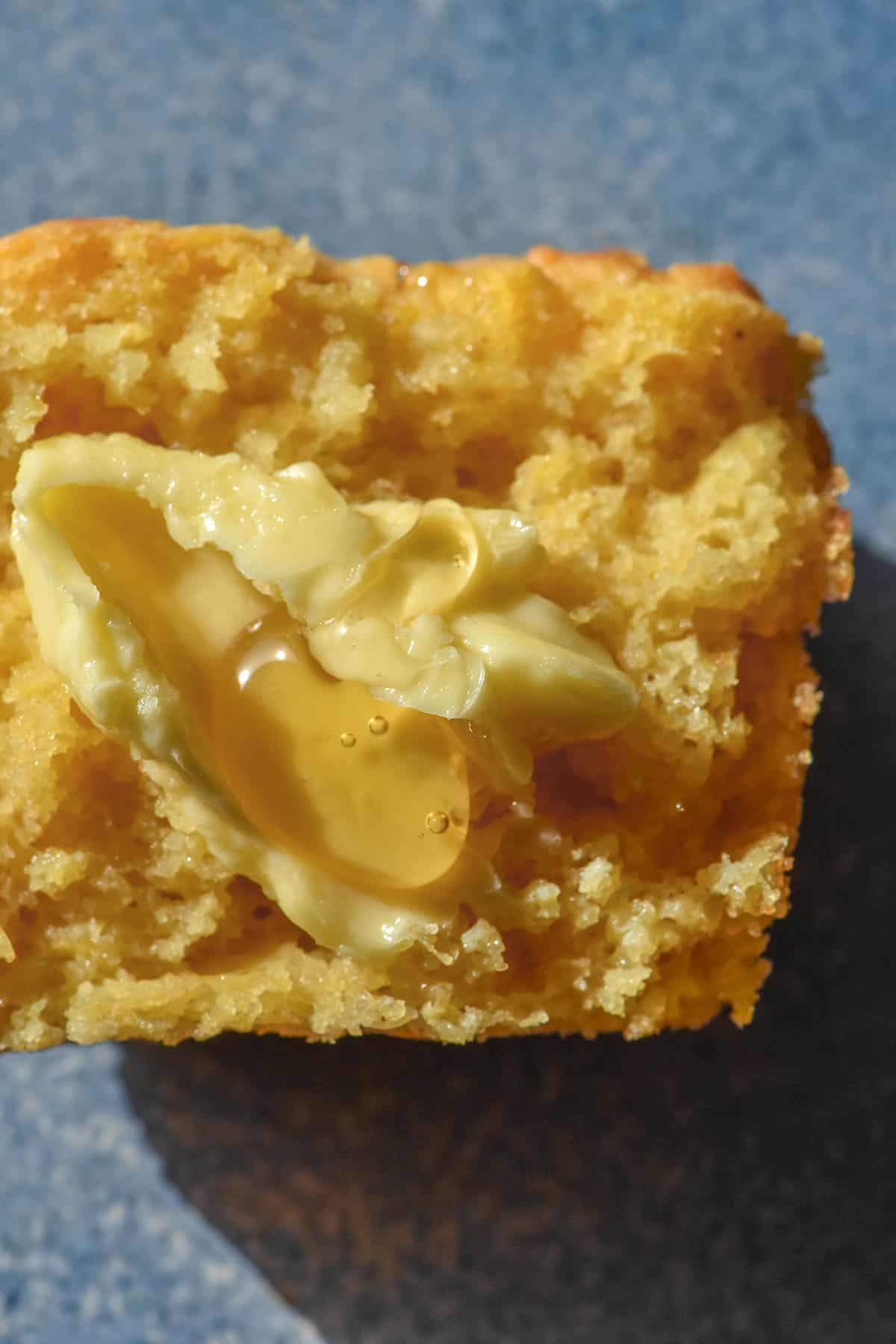 An aerial macro close up of a gluten free cornbread muffin half on a bright blue ceramic plate. The muffin half is slathered in butter and drizzled with honey. 