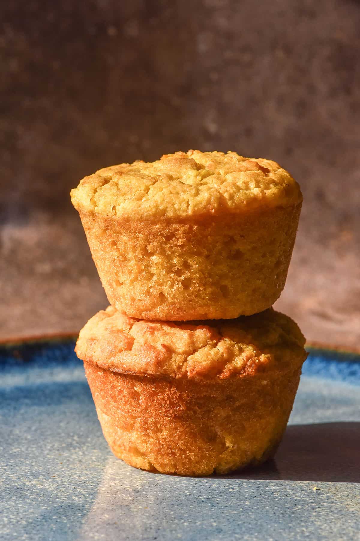 A side on image of two gluten free egg free cornbread muffins stacked on top of each other on a bright blue ceramic plate against a dark brown backdrop