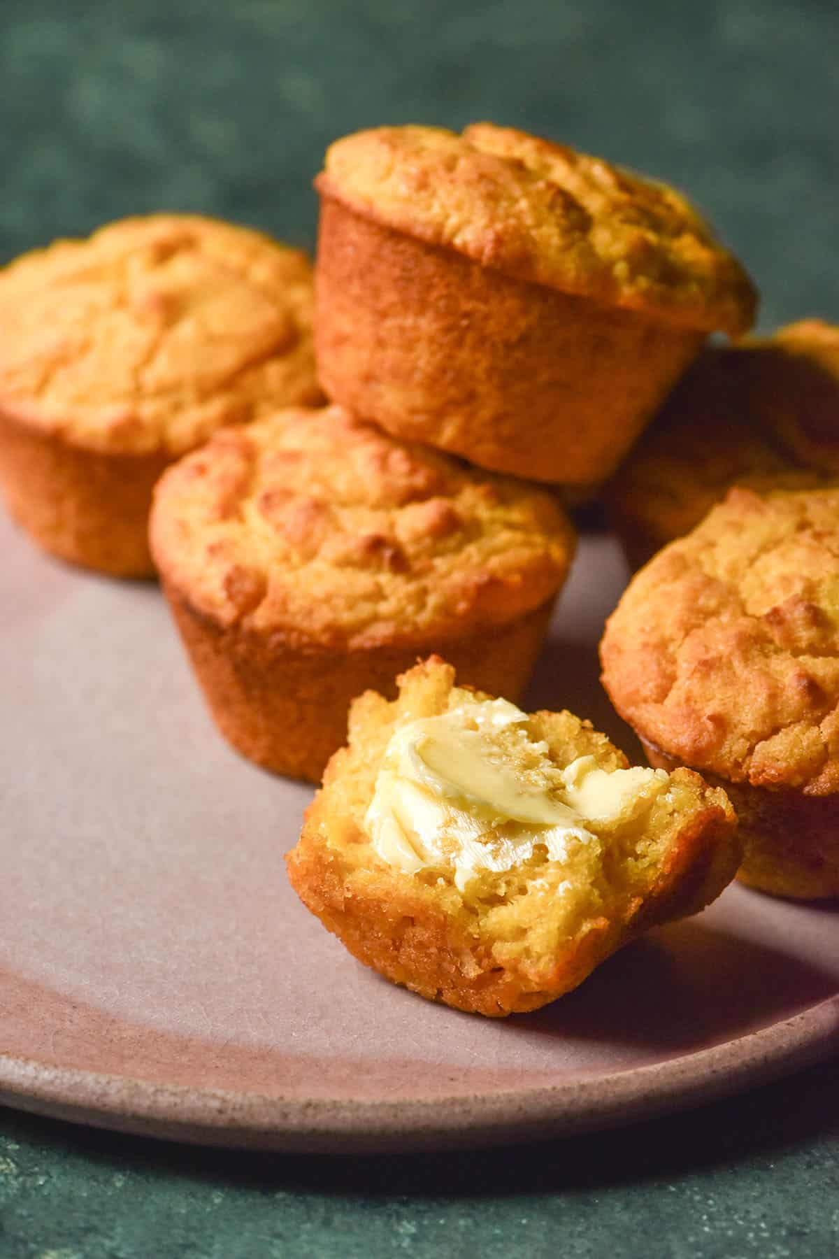 A side on image of gluten free cornbread muffins casually arranged on a pale pink plate atop an olive green backdrop. One muffin has been split in half and slathered with butter.