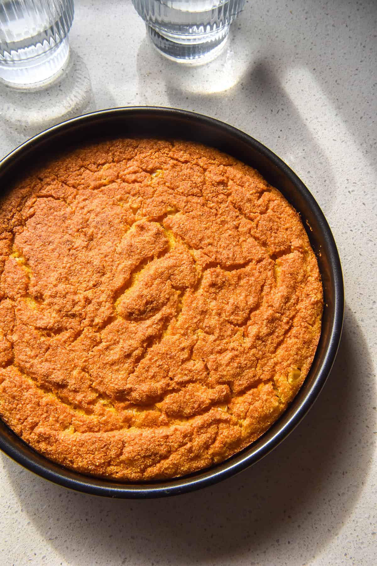 A brightly lit aerial image of a round gluten free vegan cornbread on a white countertop in bright sunlight. Two sunlit glasses of water sit in the top of the image