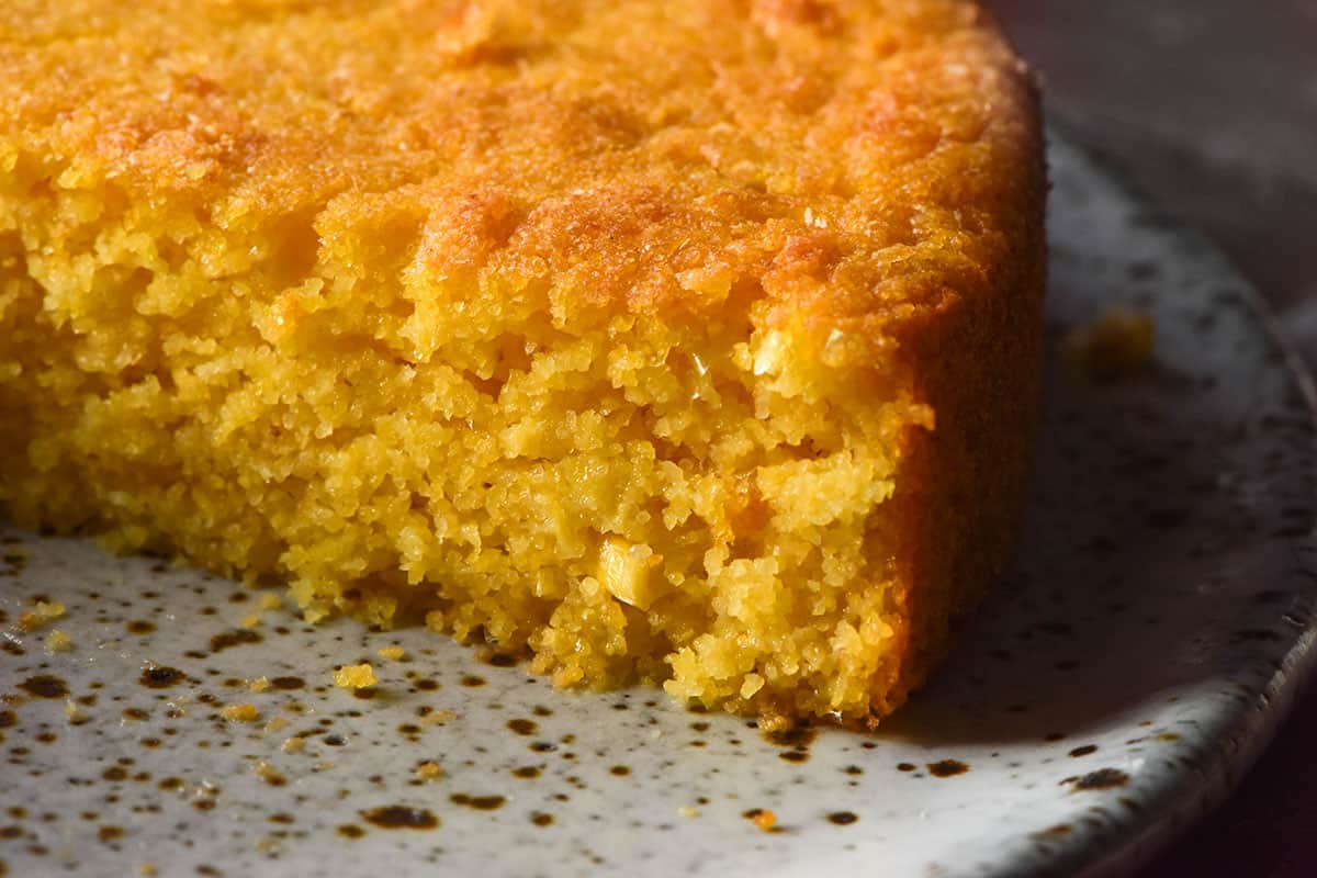 A side on close up image of a gluten free vegan cornbread atop a white speckled ceramic plate. The cornbread has been sliced to reveal the soft inner crumb. 