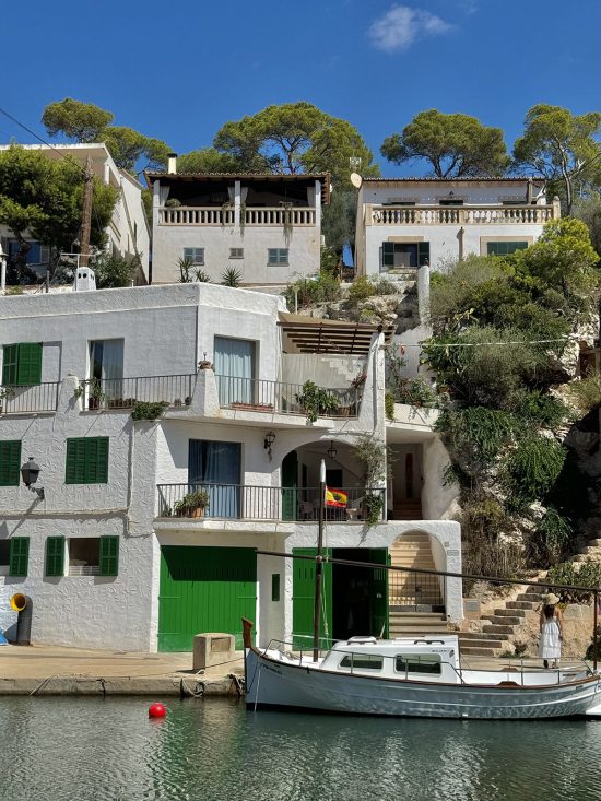 An image of the houses and water at Cala Figuera, Mallorca. The houses are set against a sunny sky and a boat sits in the water in front of the house.