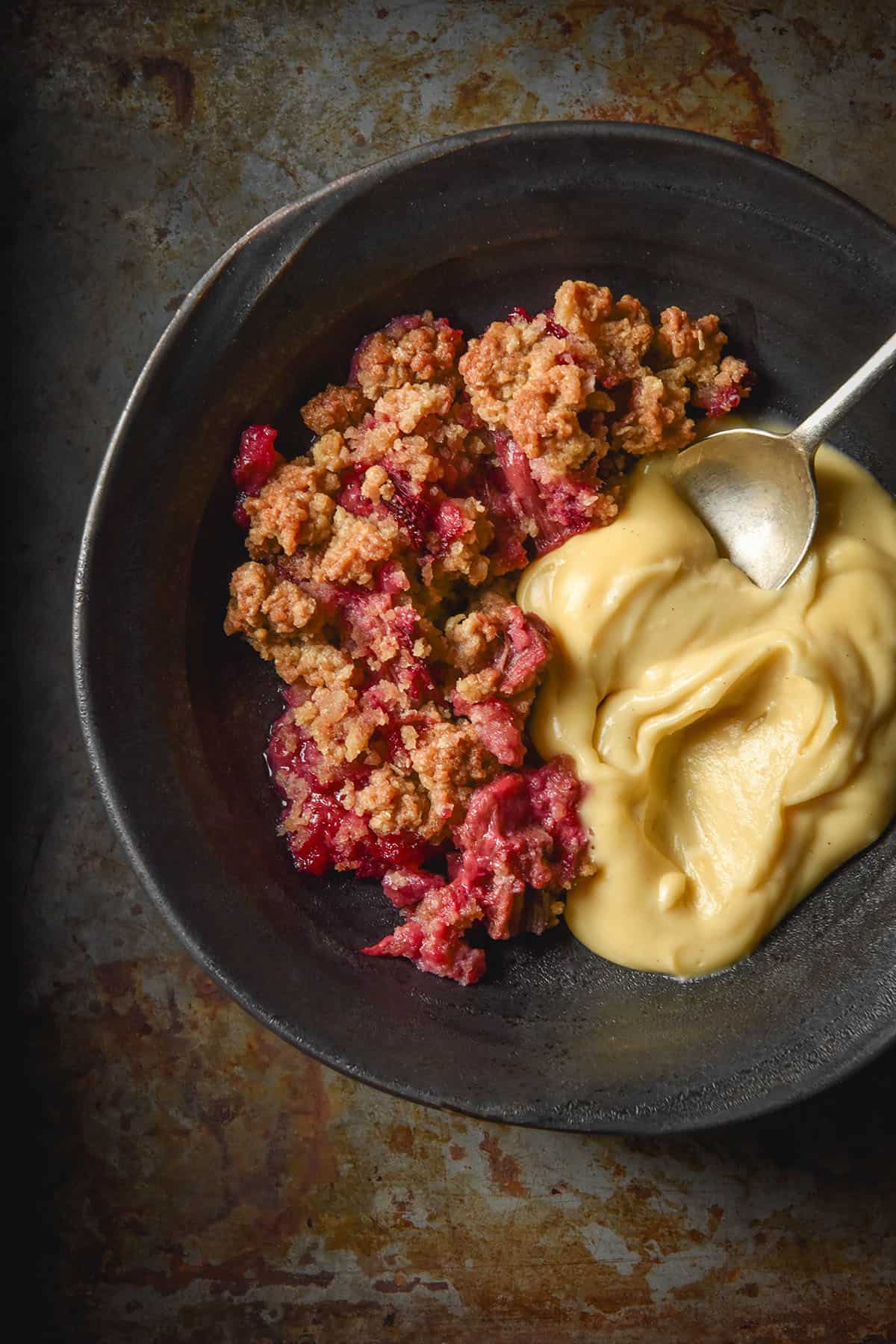 An aerial image of a dark blue ceramic bowl filled with gluten free rhubarb crumble and dairy free custard. The bowl sits atop a dark grey steel backdrop. 