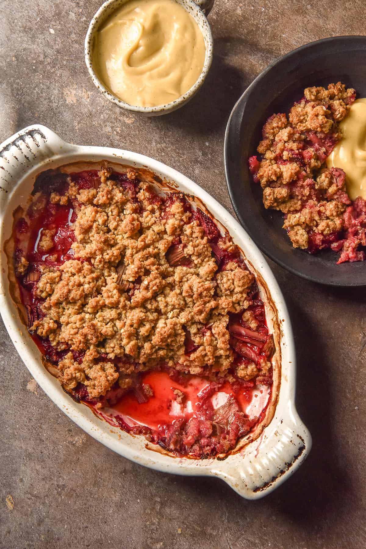 An aerial image of a white oval ceramic dish filled with gluten free rhubarb crumble. The dish sits on a dark brown backdrop and is surrounded by a water glass, a ceramic dish filled with dairy free custard and a bowl filled with crumble and custard