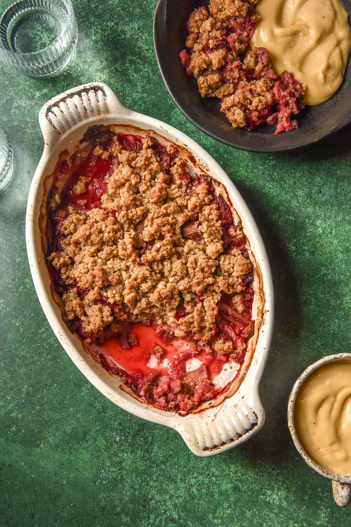 An aerial image of a gluten free rhubarb strawberry crumble in a white oval dish on an olive green backdrop. The dish is surrounded by water glasses, a ceramic dish filled with dairy free custard and a plate of crumble and custard.