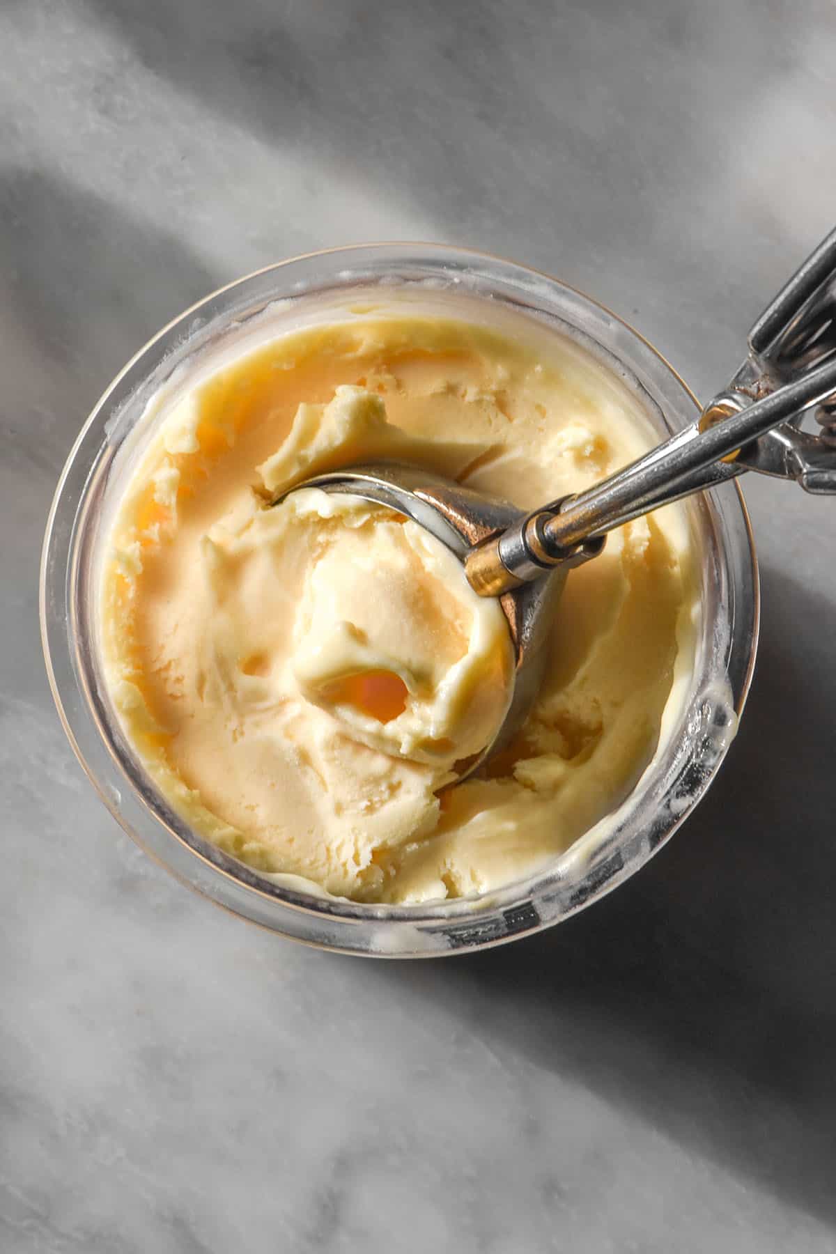 An aerial image of a tub of Ninja Creami lemon ice cream on a white marble table with a steel scoop sticking out from the tub