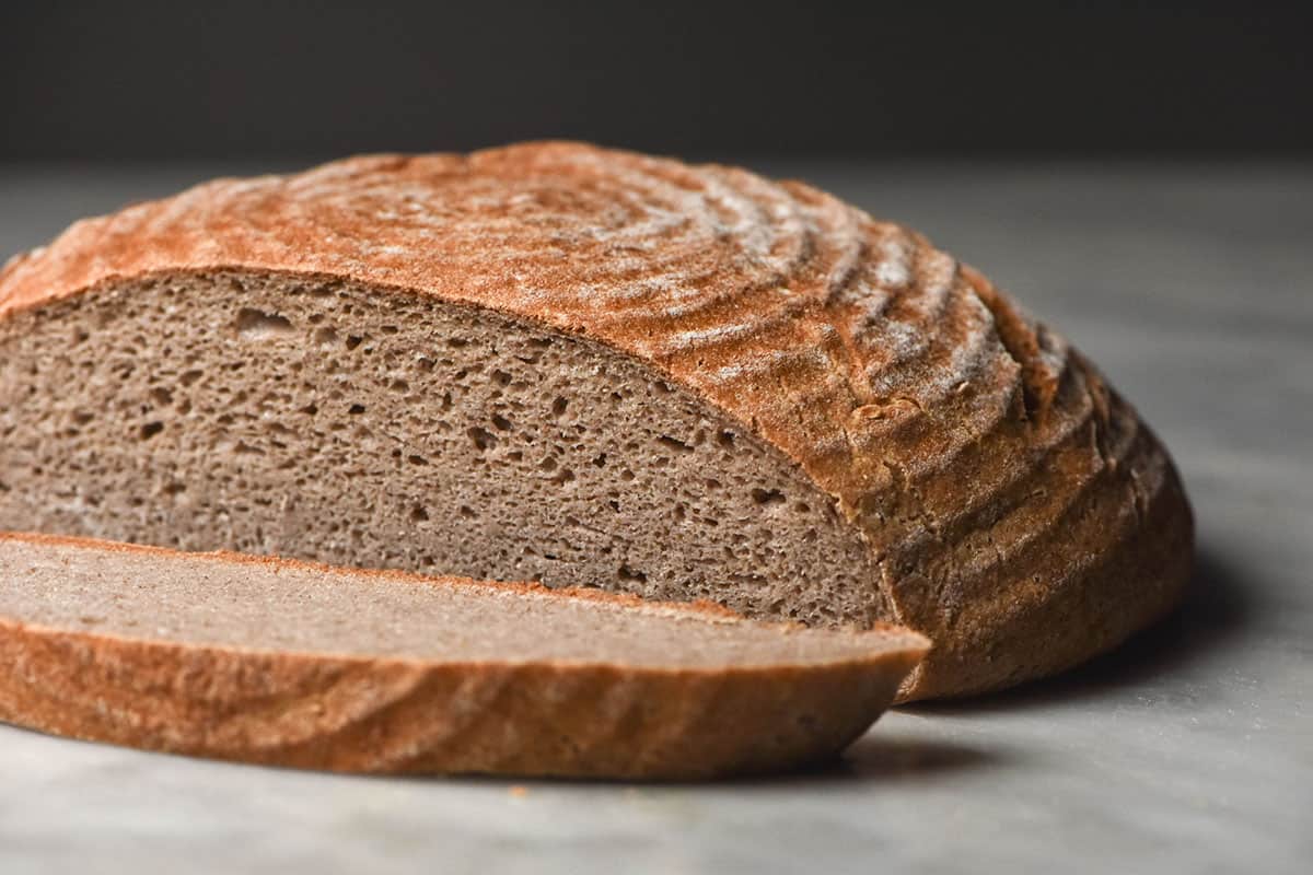 A side on macro image of a gluten free buckwheat boule that has been sliced, revealing the crumb inside. The bread sits on a white marble table against a dark backdrop