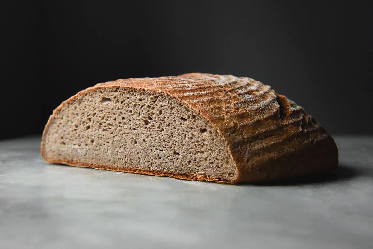 A side on image of a gluten free buckwheat boule that has been sliced to reveal the crumb of the bread. The boule sits on a white marble table against a dark backdrop