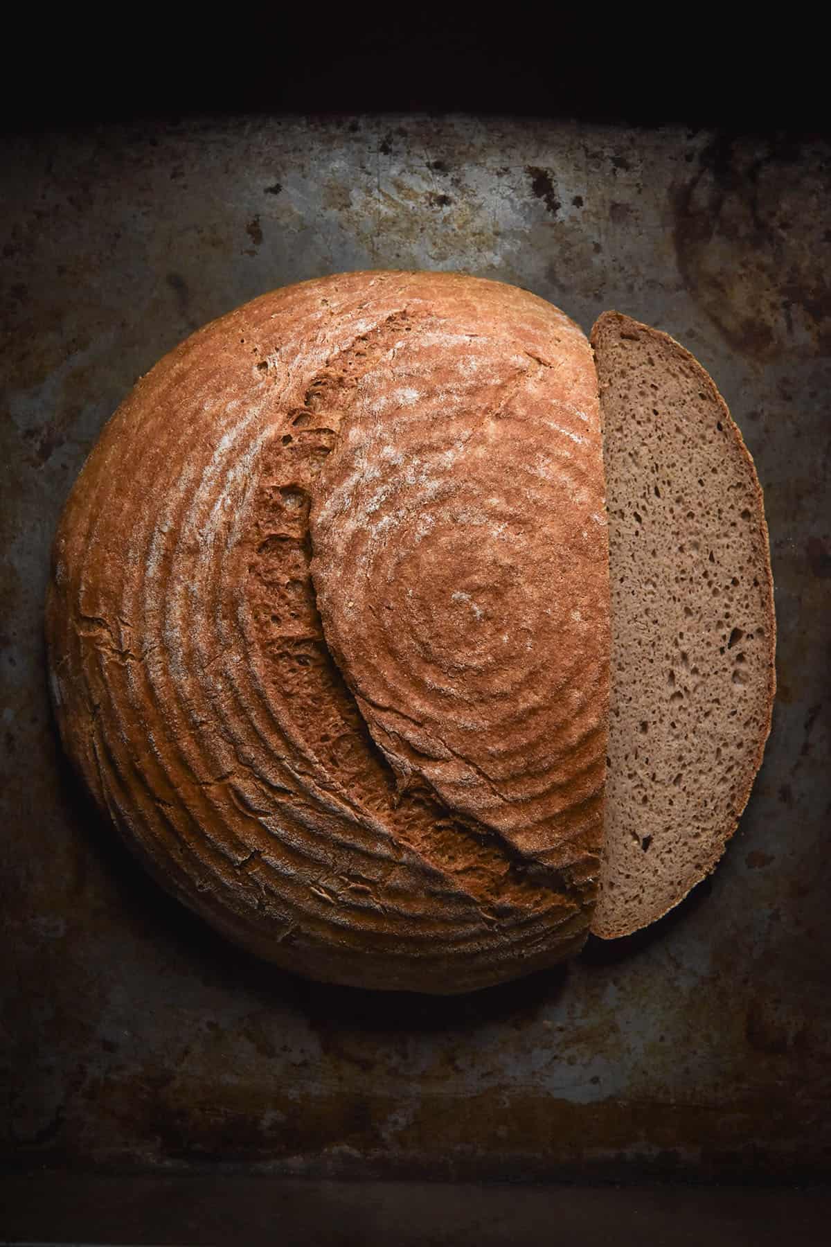 An aerial image of a gluten free buckwheat boule on a dark grey steel backdrop. The loaf has been sliced and the slice sits on the right side of the bread.